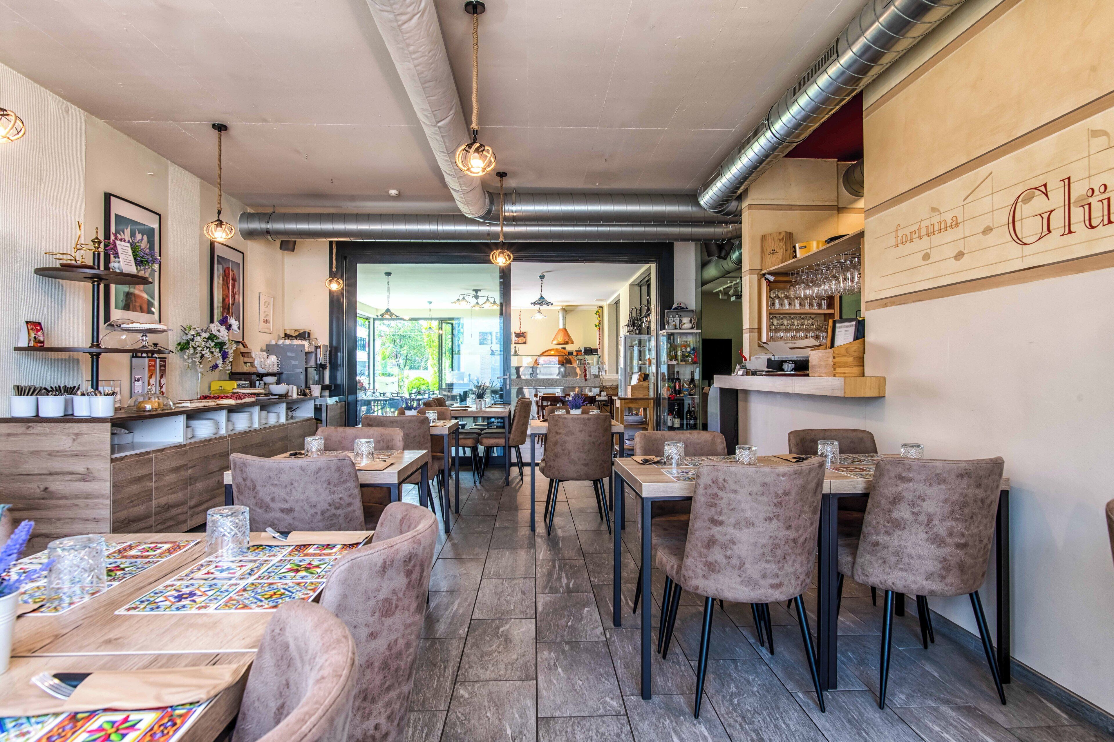 A restaurant interior with multiple dining tables and chairs, a kitchen area in the background, and a sign on the wall.