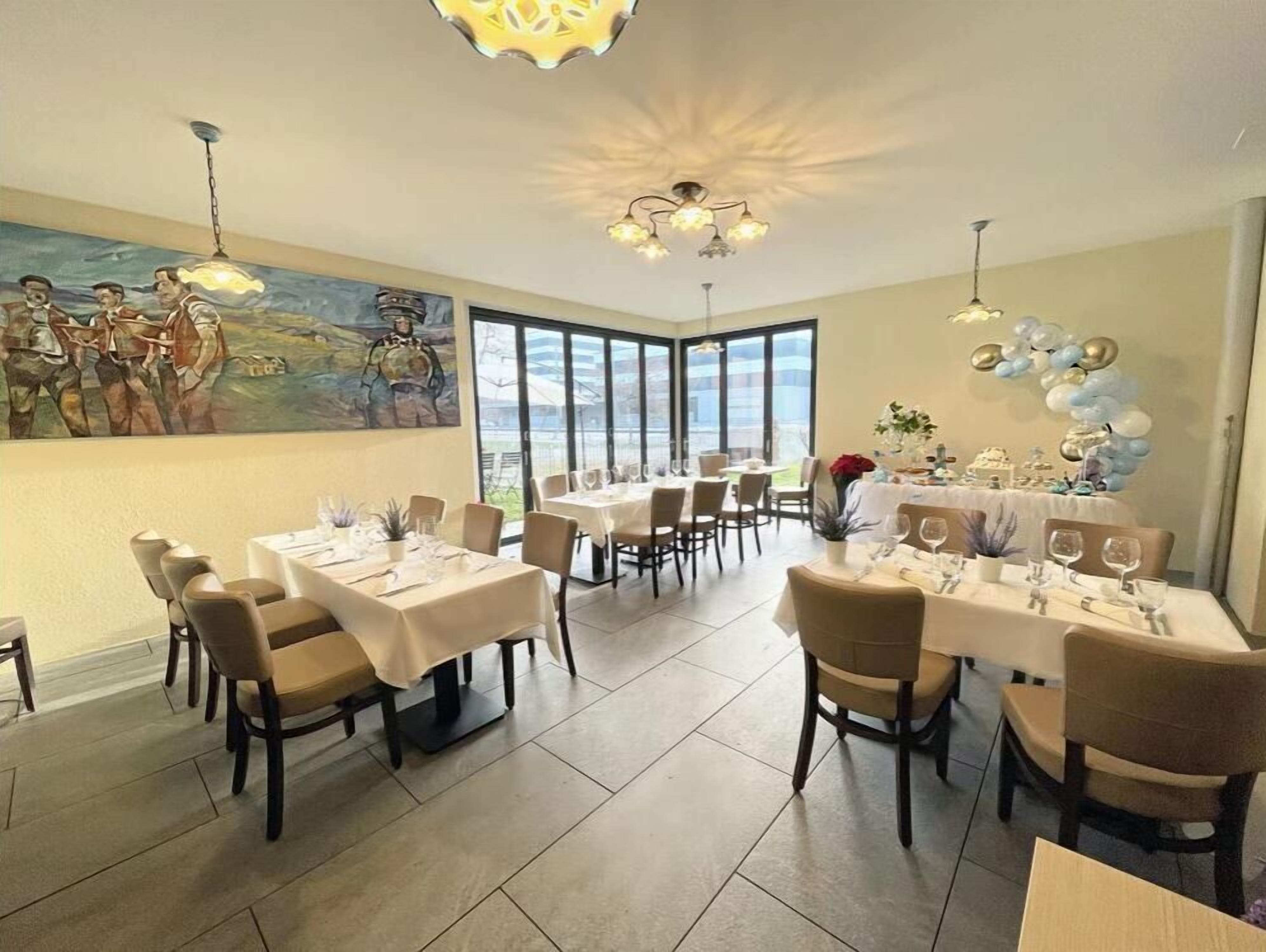 Un espace repas avec plusieurs tables dressées pour les clients, de grandes fenêtres et un plafond agrémenté d’un éclairage décoratif.