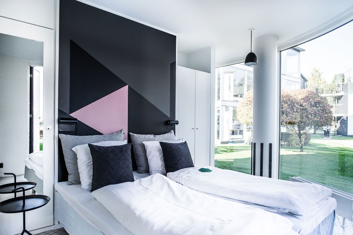 Un dormitorio moderno con una cama grande, un diseño geométrico en la pared y vistas a una zona residencial.