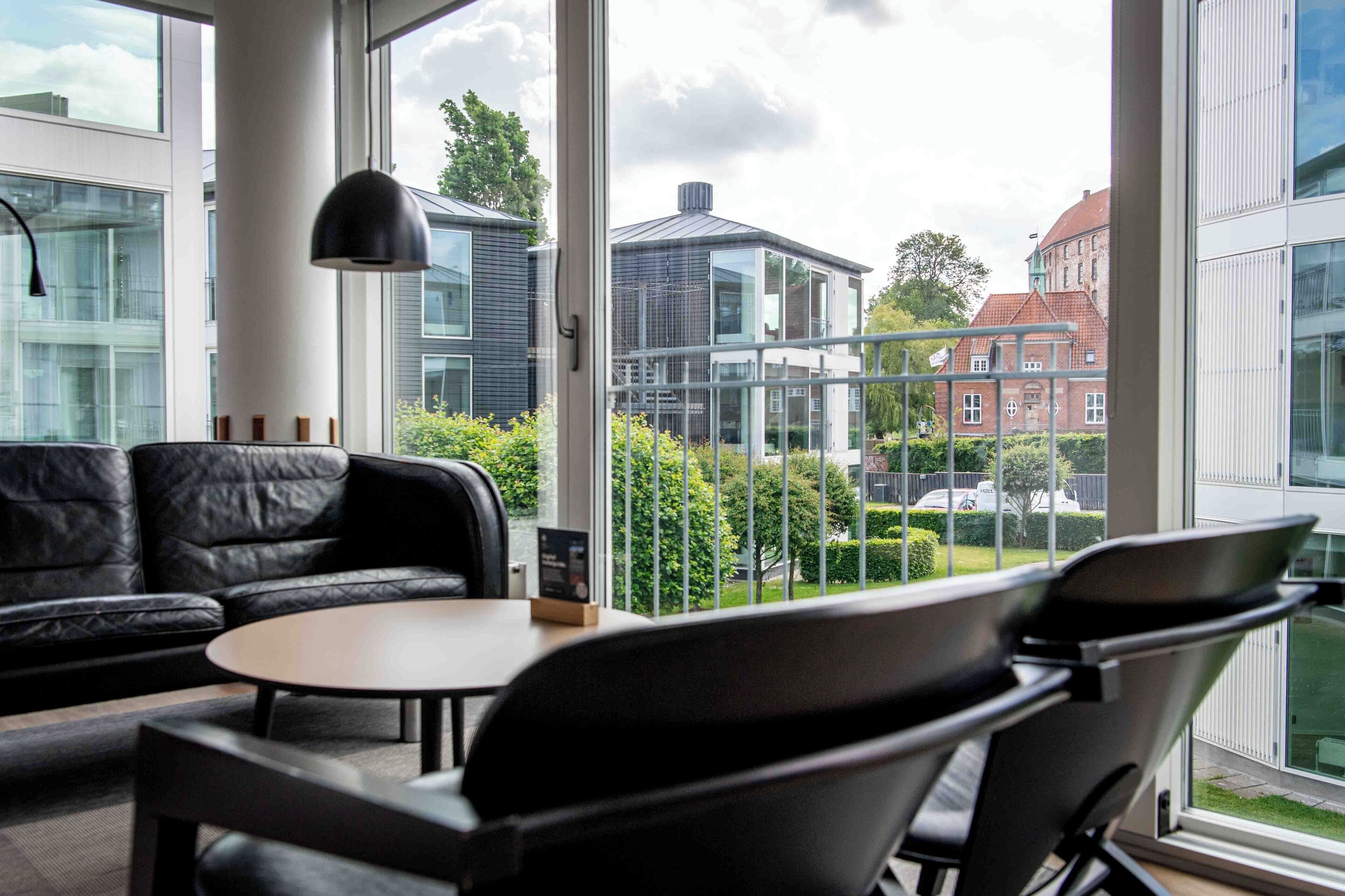 A modern interior with a leather sofa, a round table, and chairs, large windows offering a view of buildings and greenery outside.