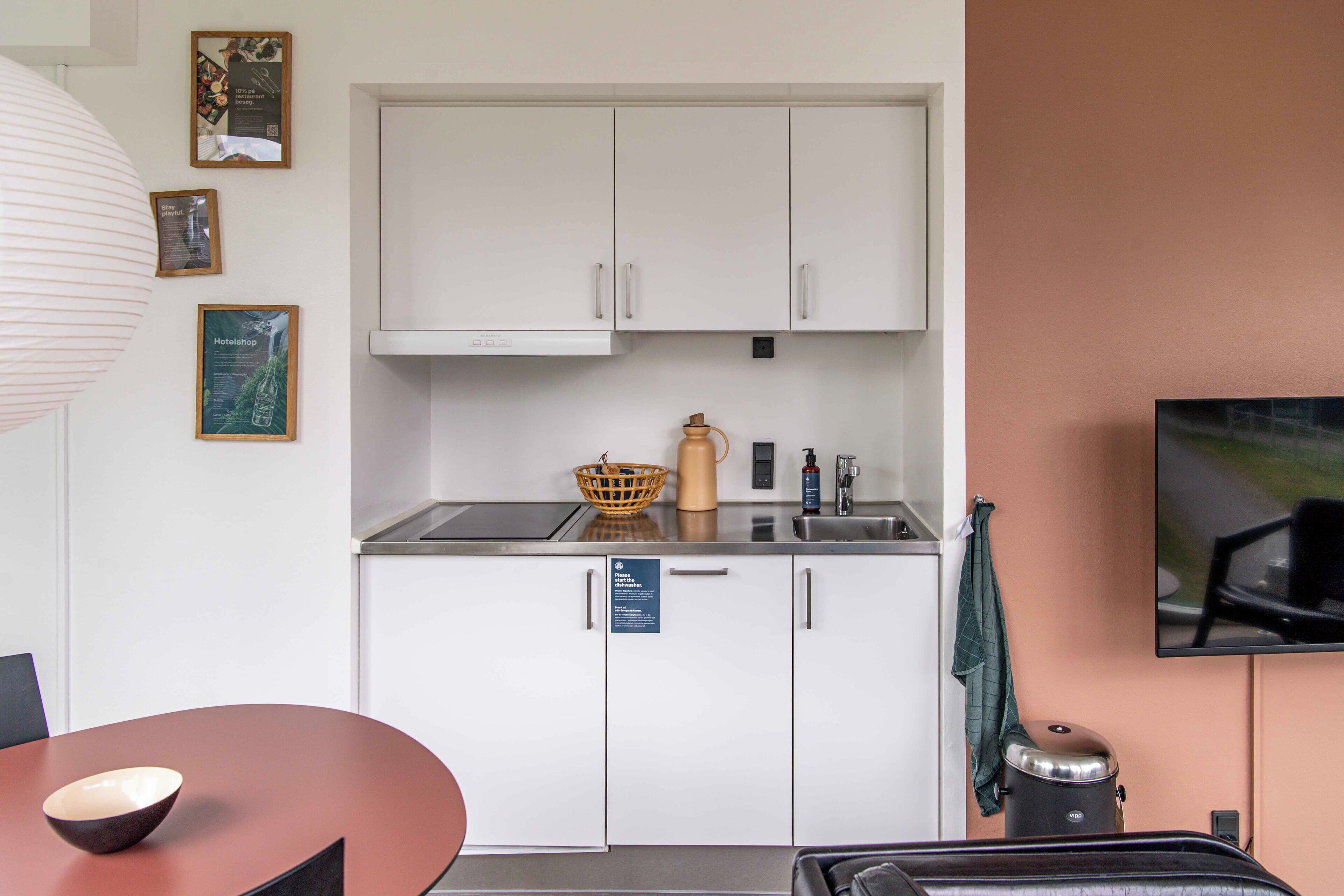 A compact kitchen with white cabinets, a sink, and a kettle. There are framed pictures on the wall and a television mounted above the counter.
