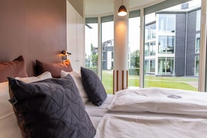 Una habitación de hotel moderna con una cama grande, varios cojines, una lámpara de noche y vistas a un edificio y zonas verdes.