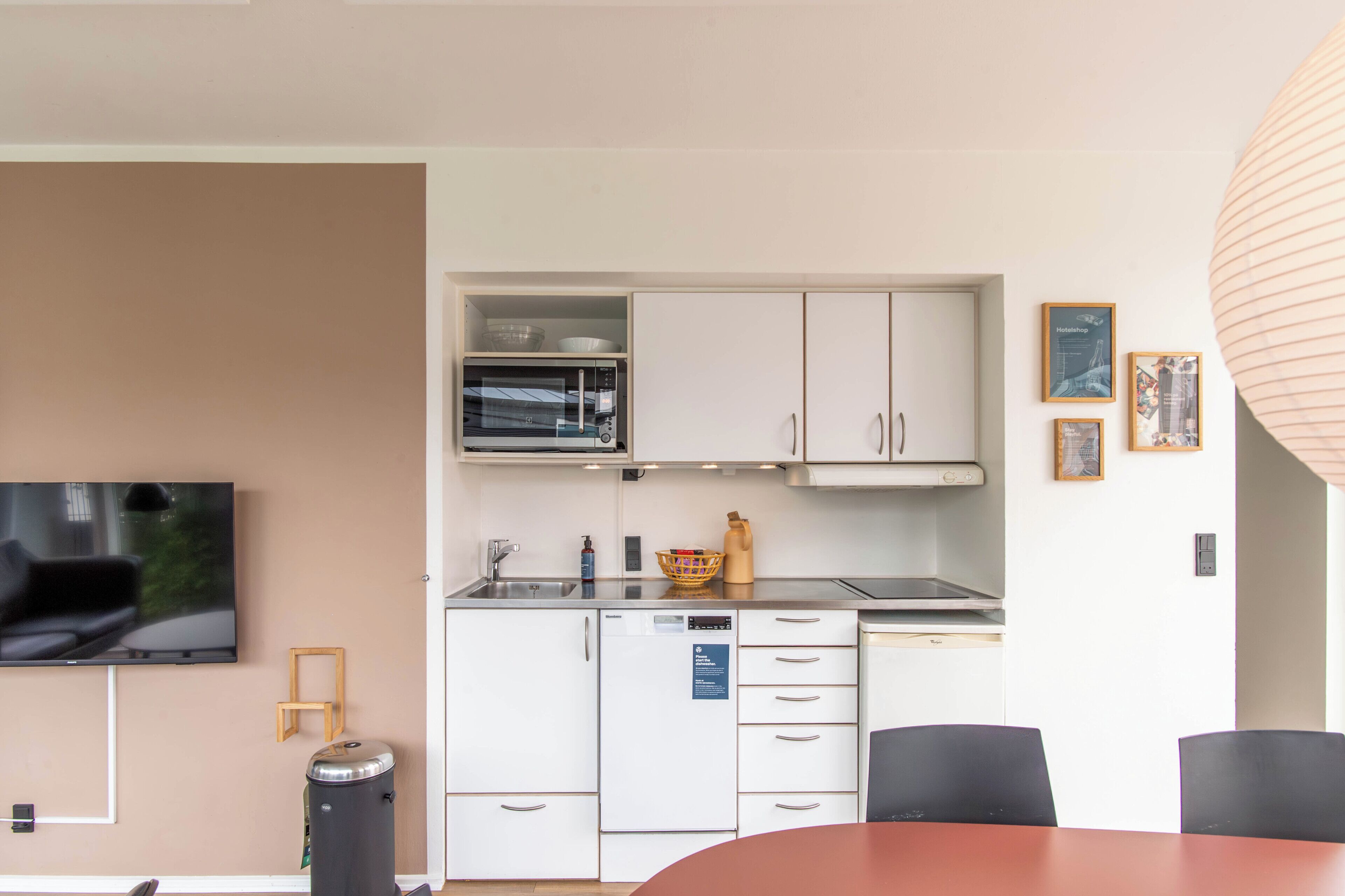 Una cocina compacta con microondas, fregadero y lavavajillas, una zona de comedor con mesa y sillas, y un televisor montado en la pared.