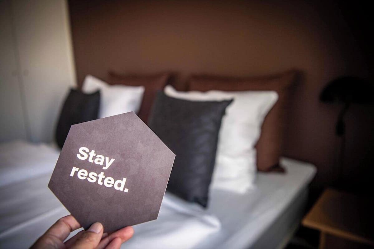 A hand holding a hexagonal card with 'Stay rested.' text, in front of a bed with pillows and a headboard.