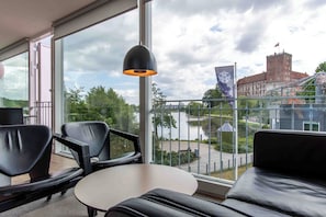 Una habitación moderna con ventanas amplias, un sofá de cuero negro, una mesa blanca y una lámpara colgante negra.