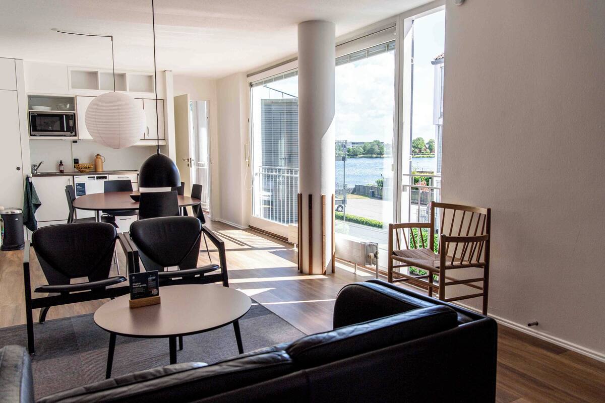 A modern living room with a sofa, a dining table with chairs, a kitchen area with a microwave, and a large window with a view of a body of water.