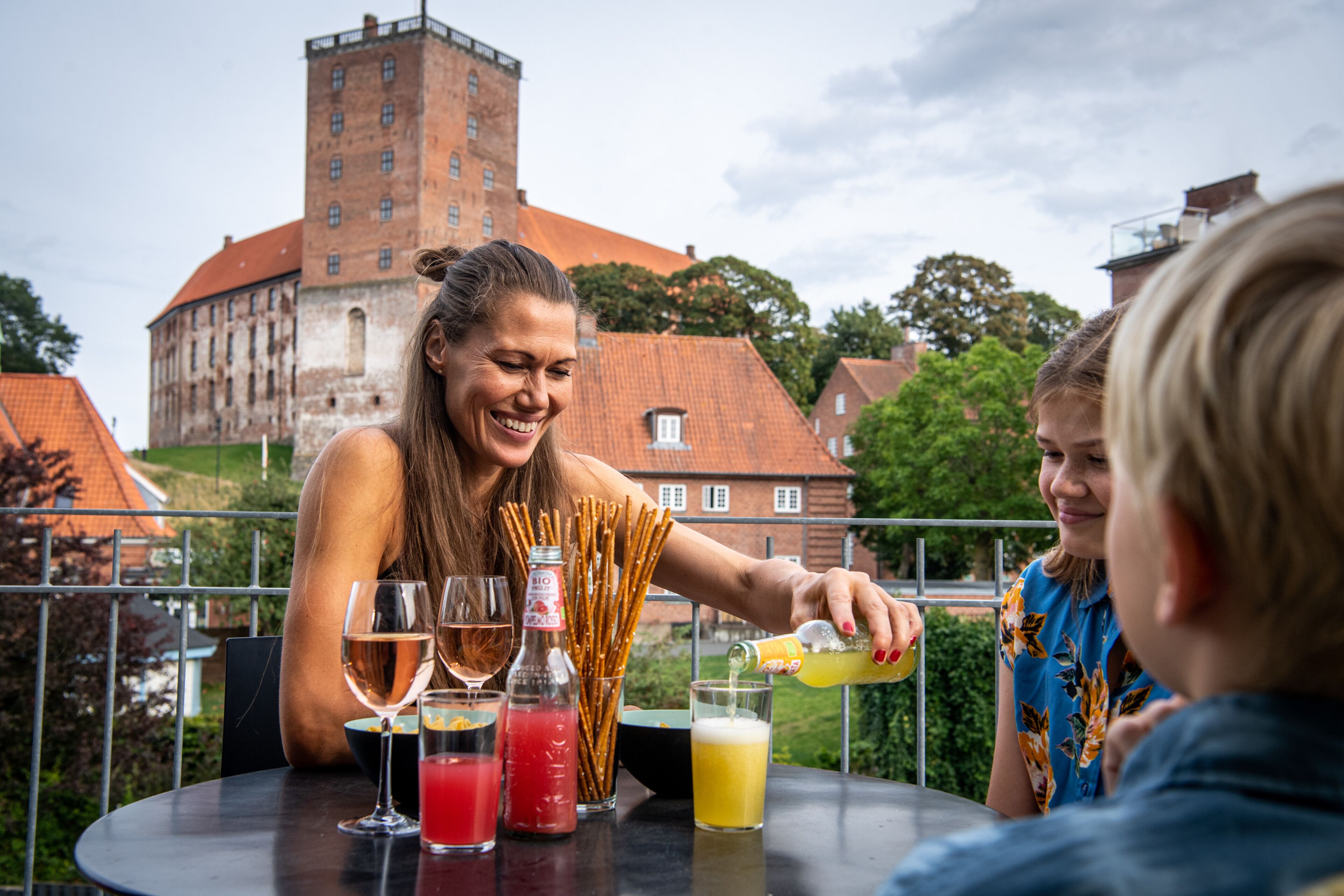 A group of people at an outdoor table with drinks and snacks, a building with a tower in the background.
