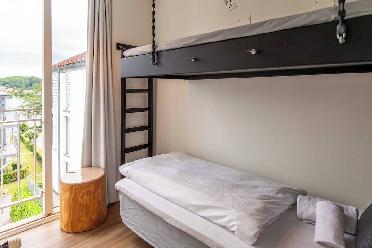 A bunk bed with a ladder, a round wooden stool, and a view of a balcony with greenery and buildings.