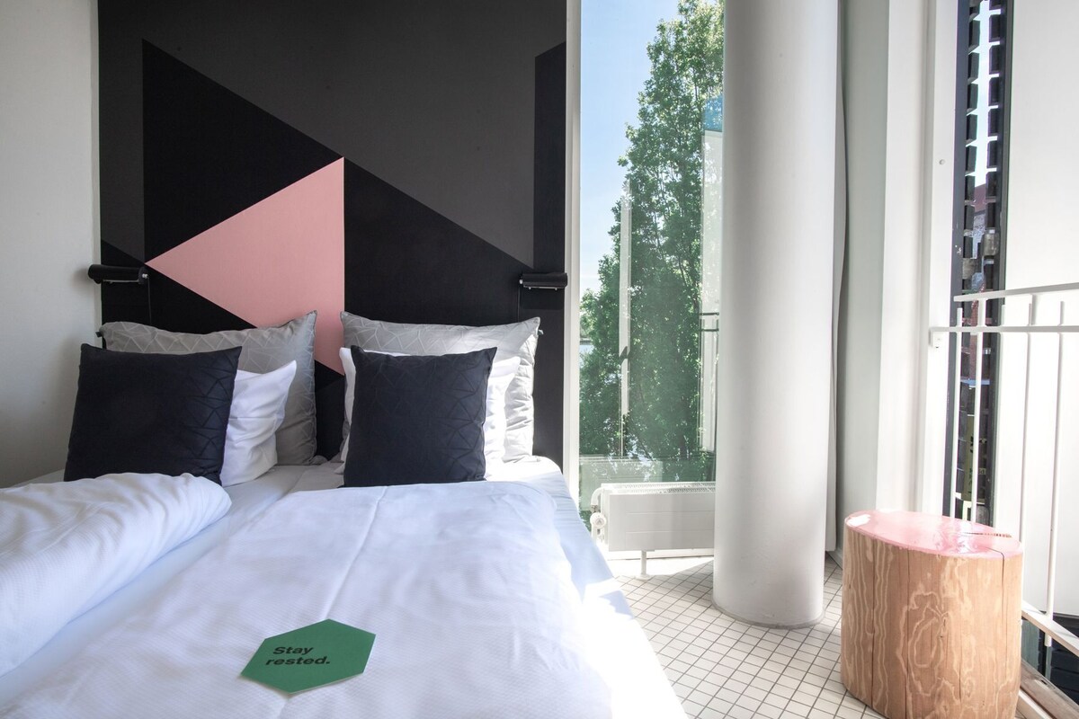 Habitación de hotel con cama, almohadas, un letrero verde, un taburete de madera y vistas a árboles a través de una ventana.