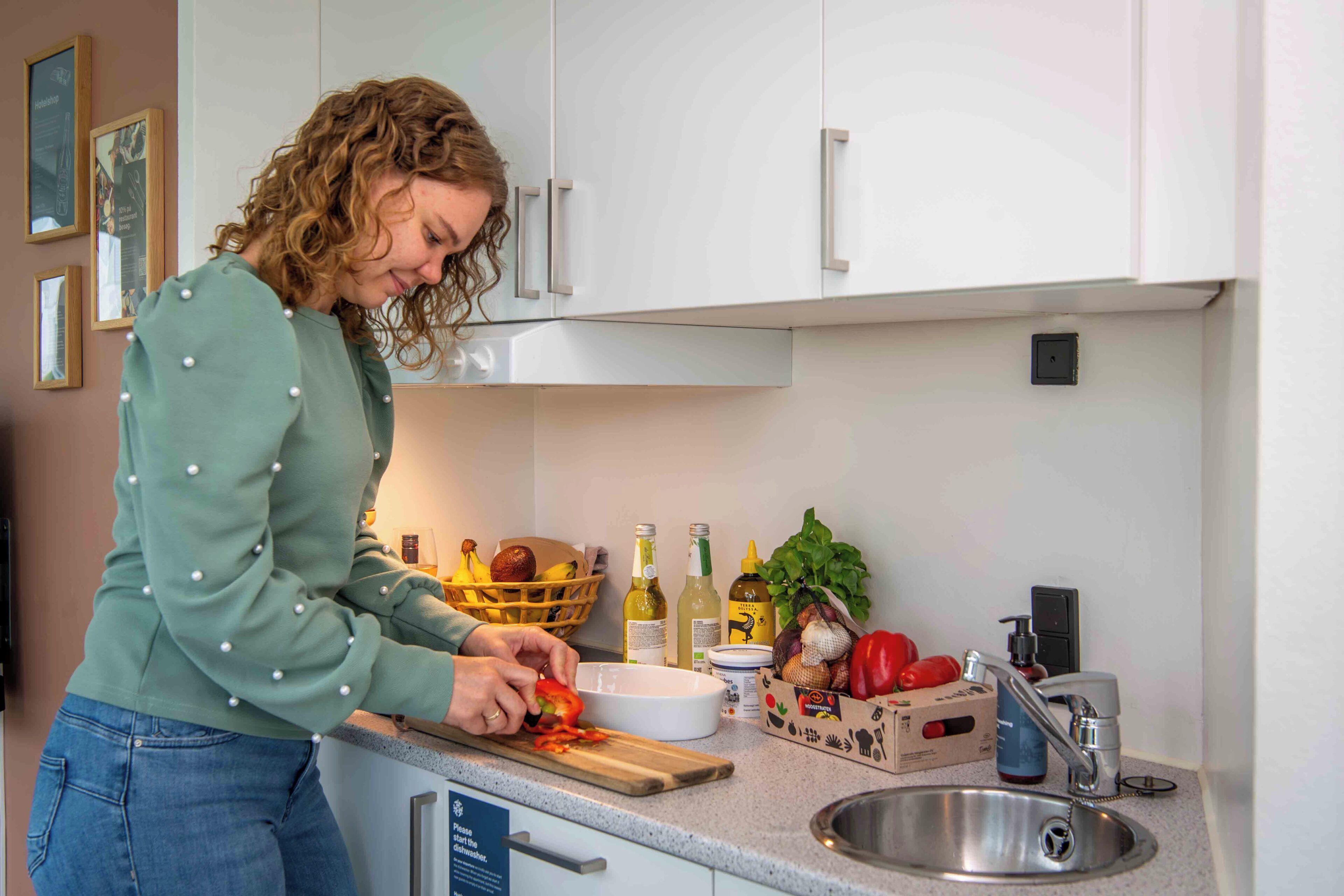 Une personne prépare des aliments dans une cuisine dotée de meubles blancs, d’un évier en acier inoxydable et de divers ingrédients disposés sur le plan de travail.