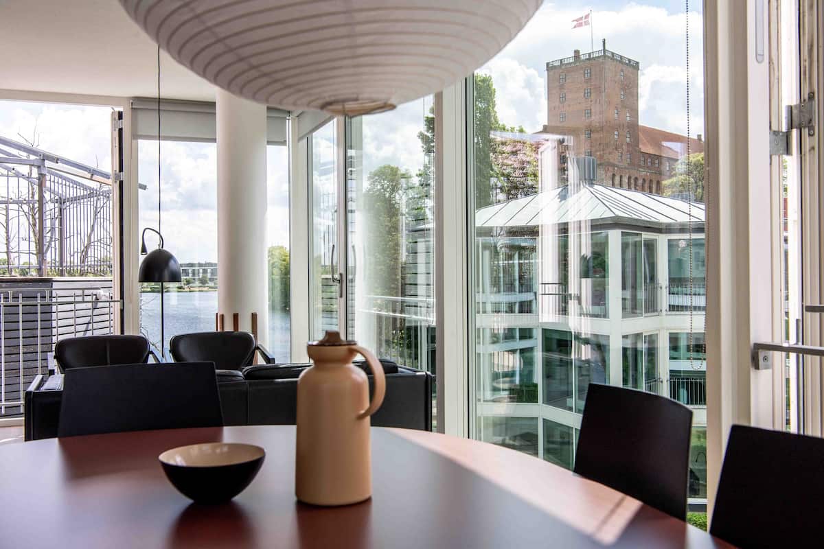 A modern dining area with a table, chairs, and a vase. Large windows offer views of a cityscape and a building with a red roof.