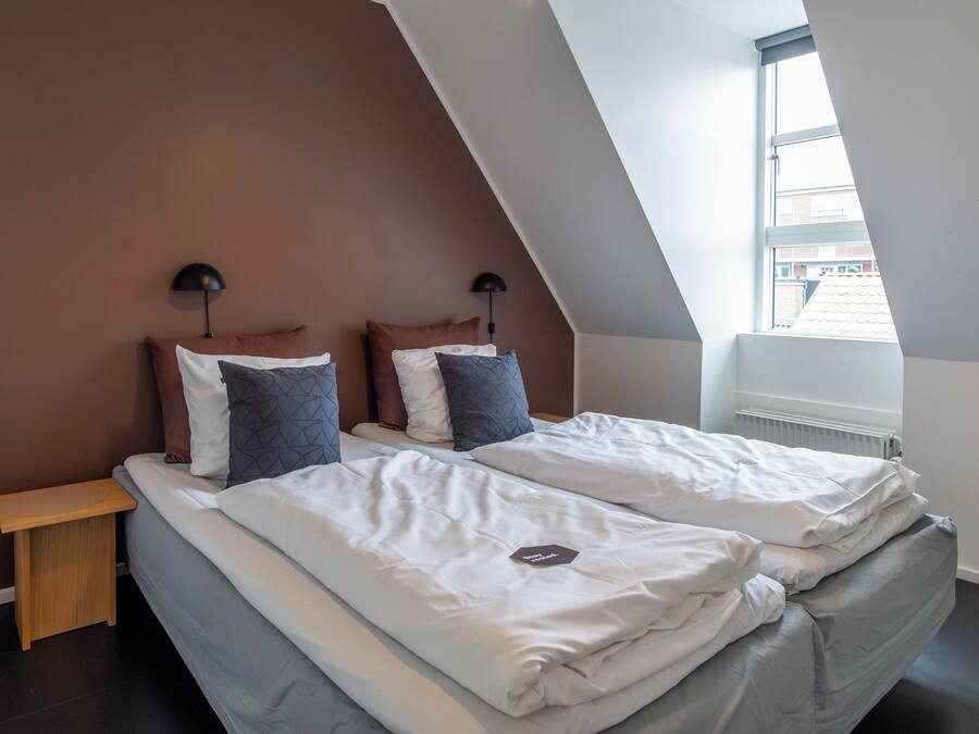 A modern bedroom with a sloped ceiling, two beds with white bedding, a wooden nightstand, and a window with a view of buildings.