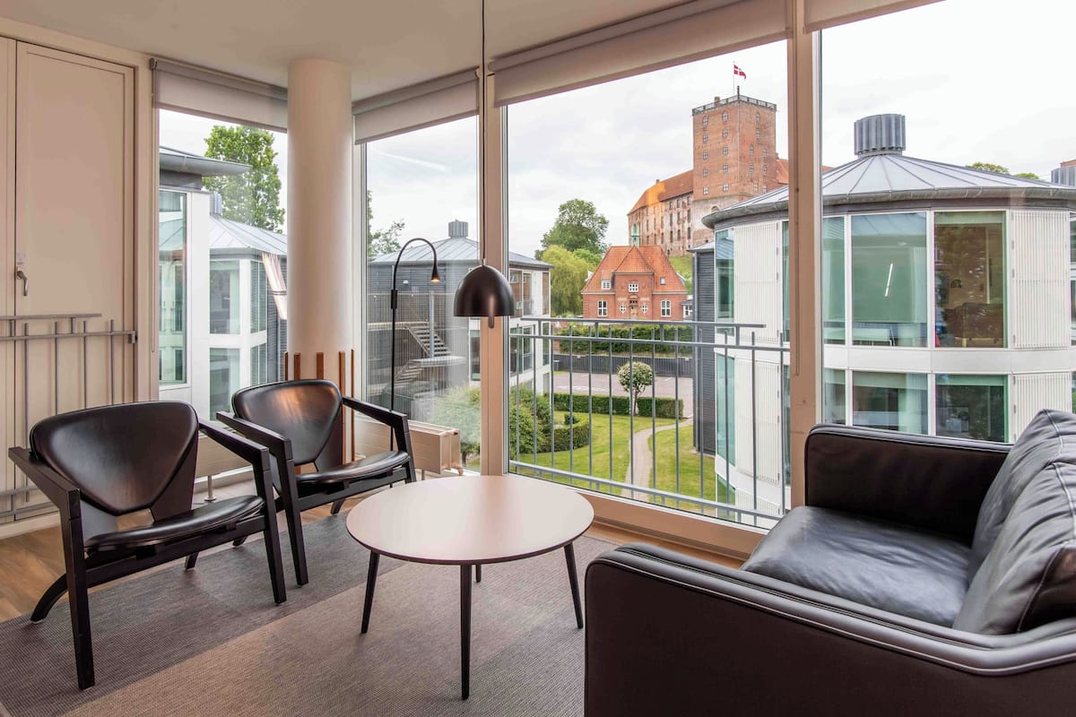 A modern room with large windows, a sofa, two chairs, a small table, and a lamp.
