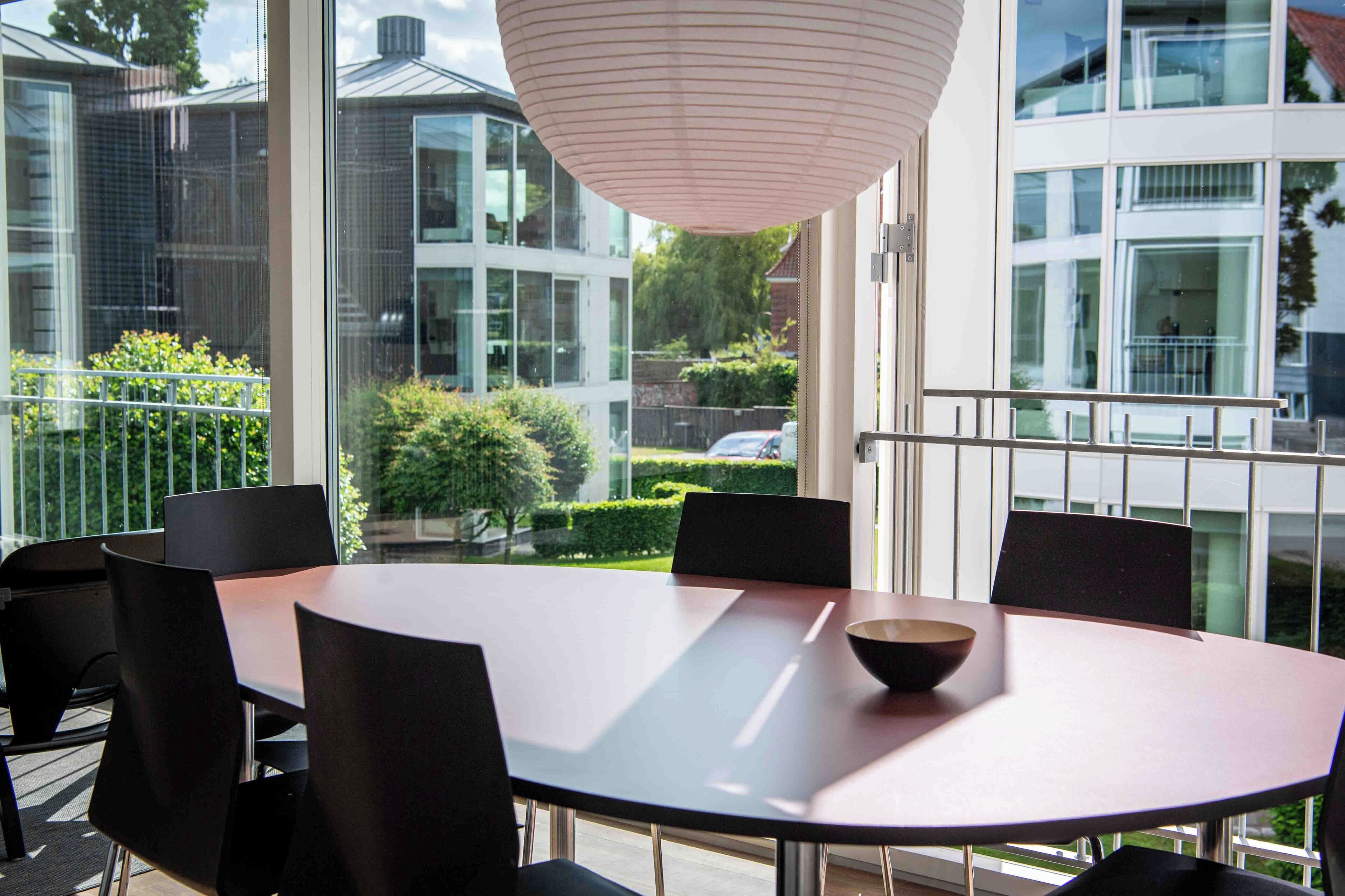 A modern dining area with a round table and black chairs, overlooking a garden and neighboring buildings.