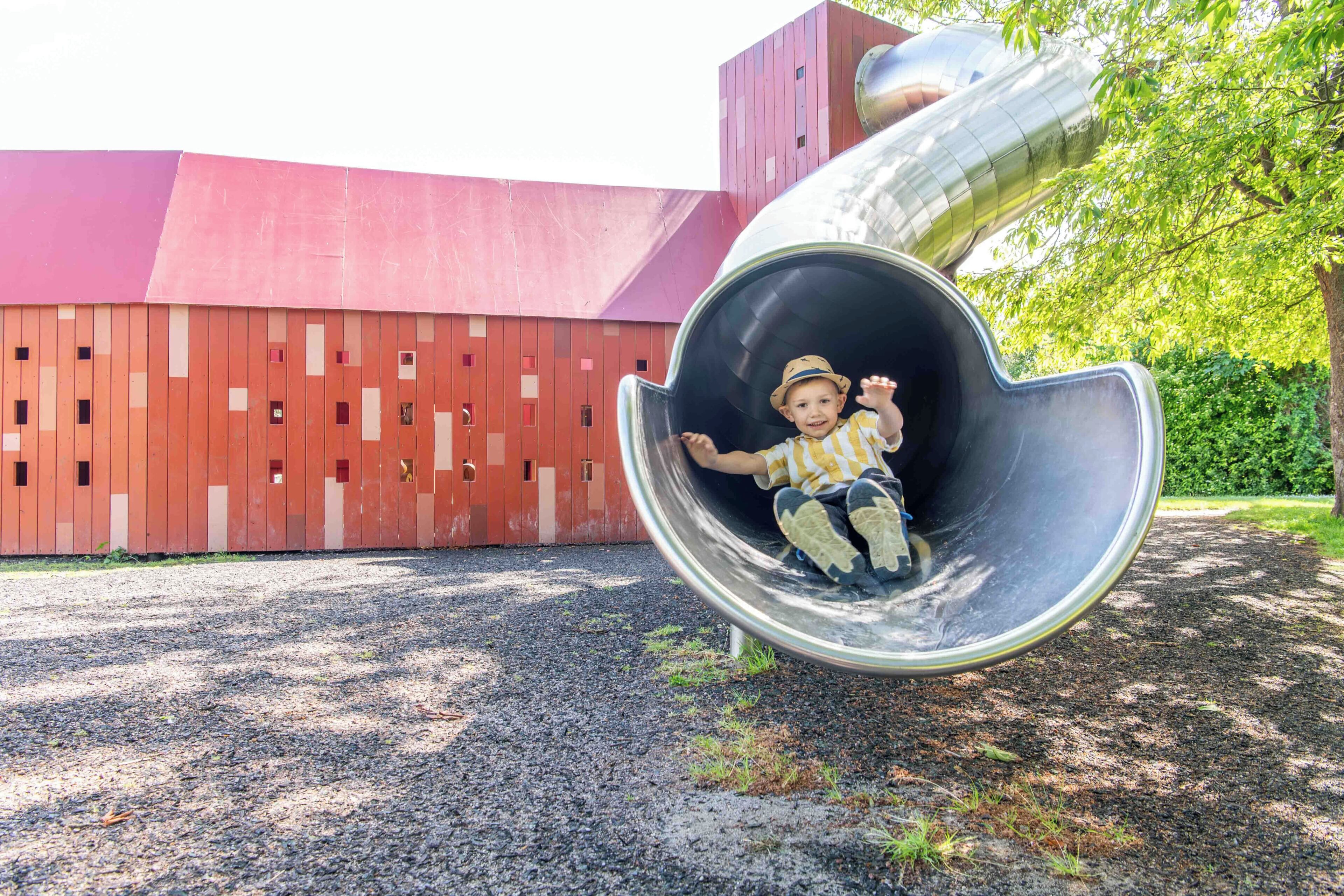 A child sliding down a large, metallic spiral slide in a playground area with a red barn-like structure in the background.