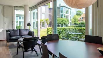 Un intérieur moderne avec un canapé, des fauteuils, une table à manger et de grandes fenêtres donnant sur des bâtiments et des espaces verts.