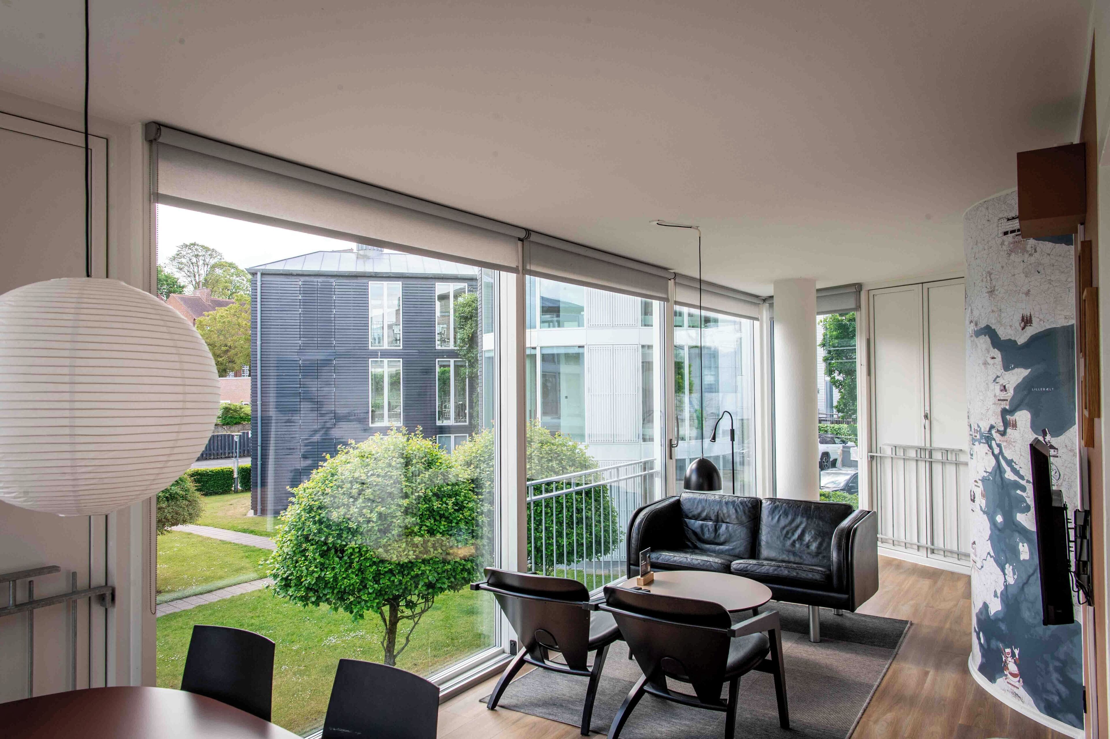 Una habitación moderna con un ventanal grande, un sofá de cuero negro, una mesa de comedor y vistas a un edificio y áreas verdes.