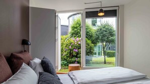A bedroom with a bed, pillows, a bedside lamp, and a view of a garden through a large window. - Kolding Hotel Apartments (Kolding)