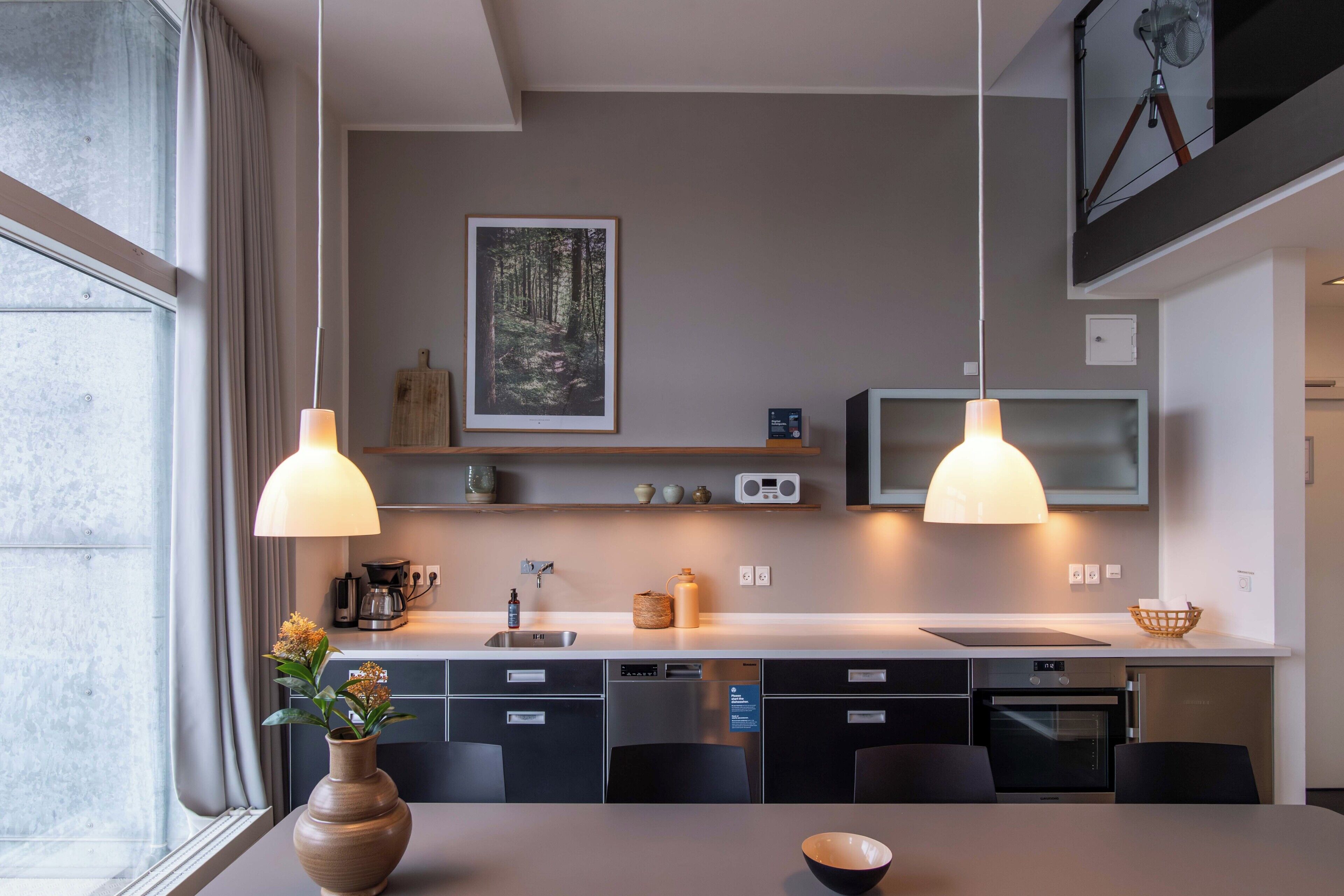 Una cocina moderna con comedor, luces colgantes, un cuadro enmarco con un paisaje forestal y electrodomésticos integrados.