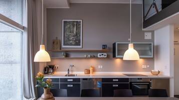 Una cocina moderna con comedor, luces colgantes, un cuadro enmarco con un paisaje forestal y electrodomésticos integrados.
