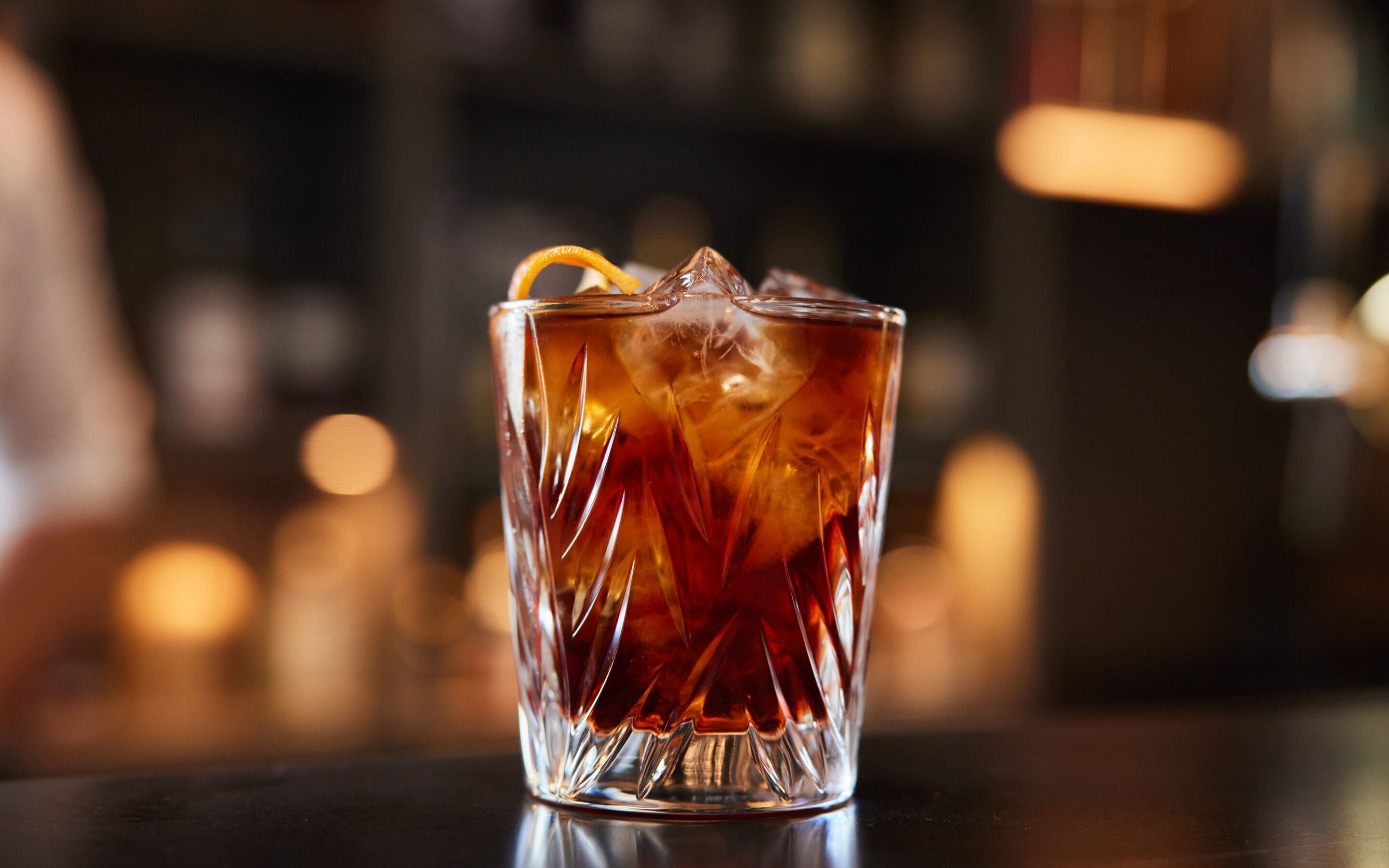 Et gennemsigtigt glas med et mønstret design, fyldt med en mørk væske og et appelsinstykke, placeret på en reflekterende overflade.