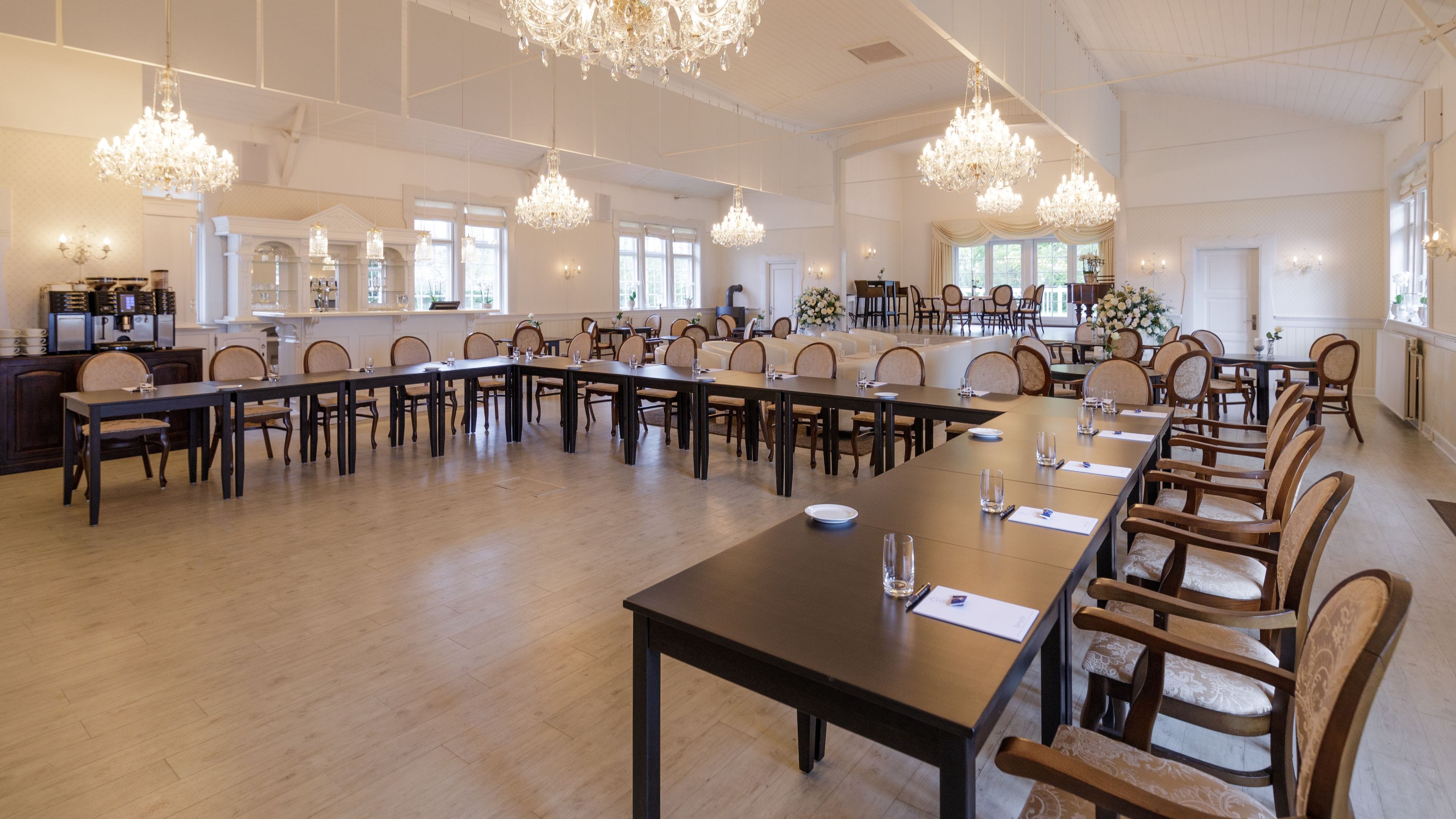Een ruime congreszaal met lange tafels en stoelen, kroonluchters en een keuken met een koffieapparaat.