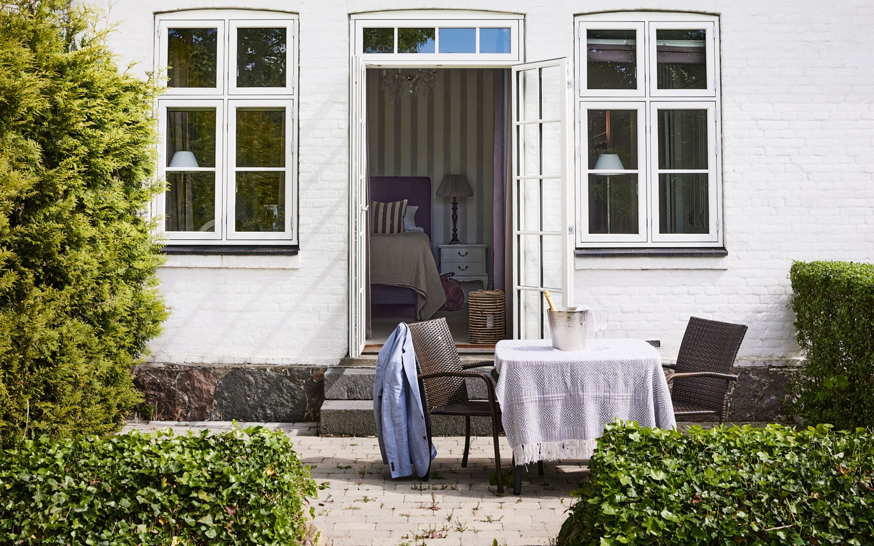 Un bâtiment blanc doté d’un patio avec jardin, comprenant une table et des chaises, un fauteuil en osier et une petite table recouverte d’une nappe.