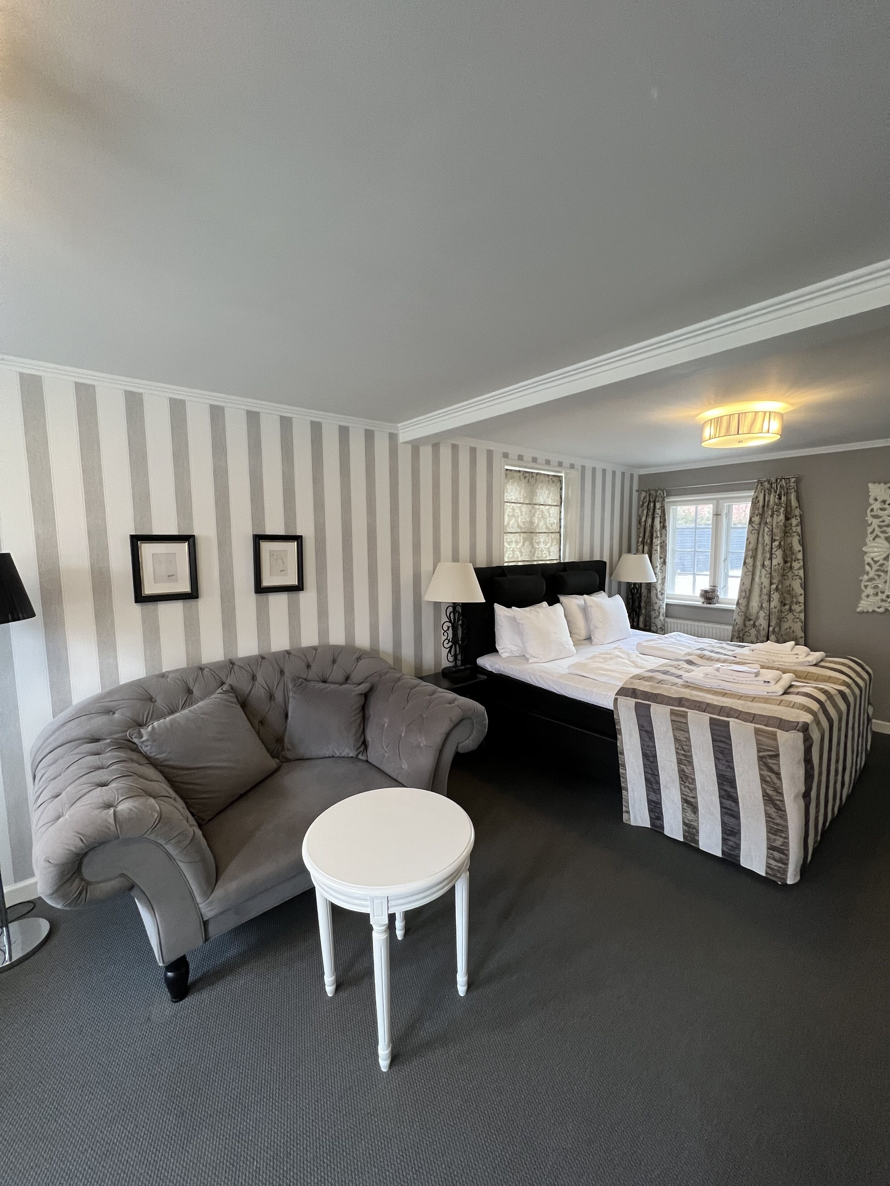 Habitación de hotel con pared a rayas grises, sofá tapizado gris, una mesa redonda blanca, cama con ropa de cama a rayas y una ventana con cortinas.