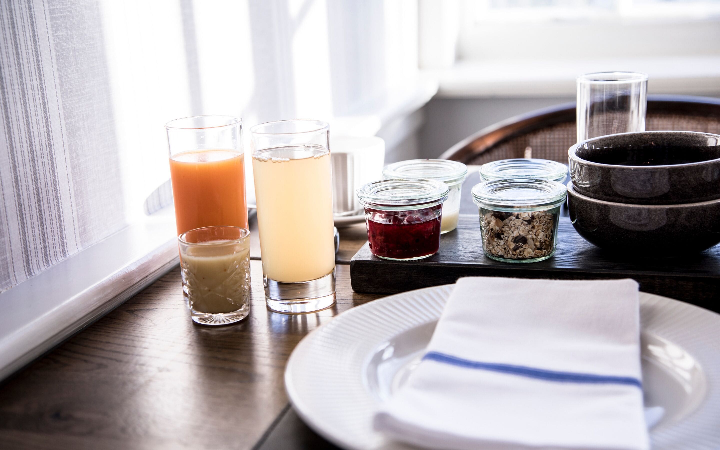 A breakfast tray with various food items and drinks, including a glass of orange juice, a glass of milk, a glass of tea, a jar of jam, a bowl of cereal, and a plate with a napkin.