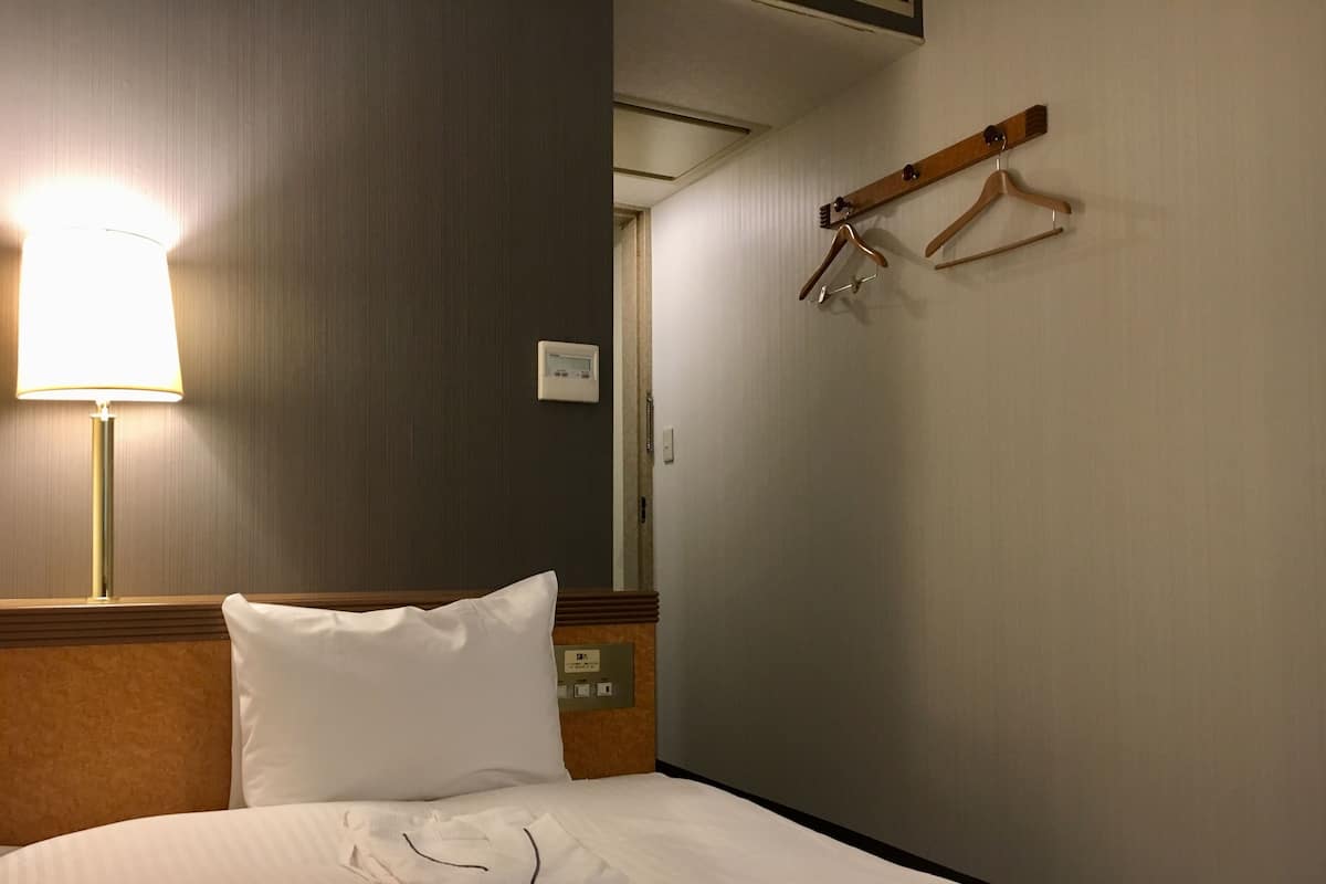 Una camera d'albergo con un letto rifatto, una lampada, un termostato e un appendiabiti con grucce.
