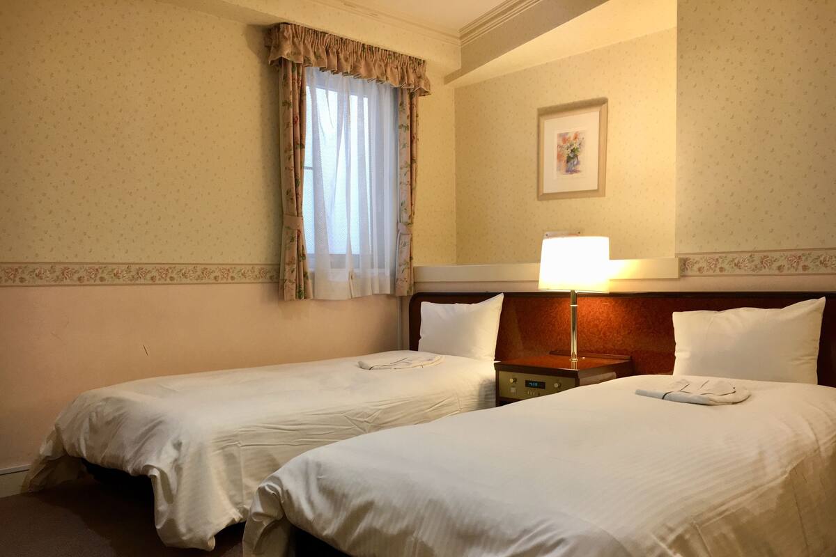 A hotel room with two single beds, a nightstand with a lamp, a window with floral curtains, and a framed picture on the wall.