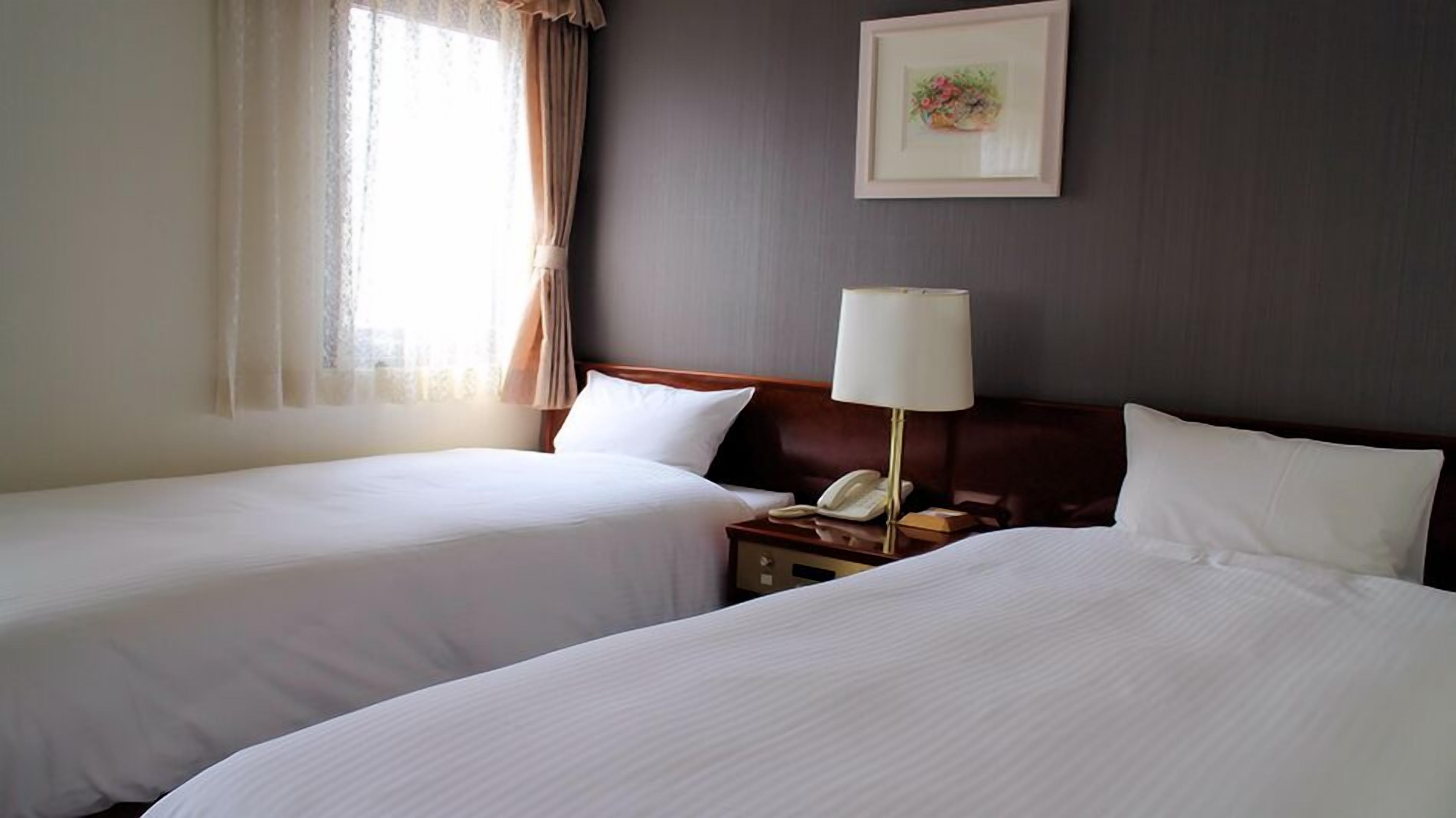 Una habitación de hotel con dos camas, una mesita de noche con lámpara, un cuadro enmarcado en la pared y una ventana con cortinas.