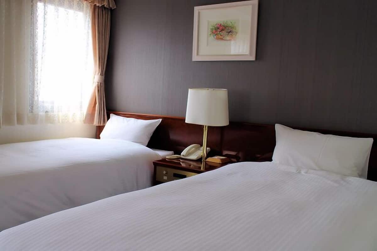 A hotel room with two beds, a bedside table with a lamp, a framed picture on the wall, and a window with curtains.