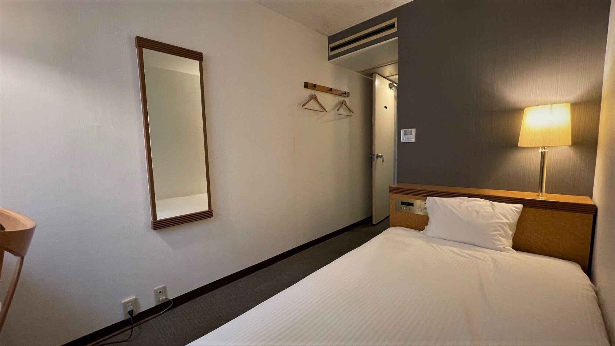 Una habitación de hotel con una cama, una lámpara de noche, un espejo y un perchero.