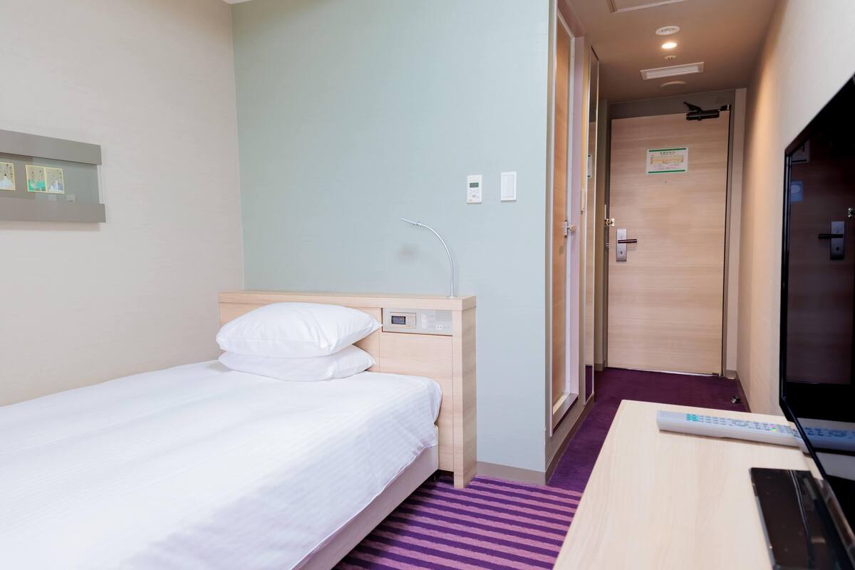 A hotel room with a single bed, a desk with a laptop, a TV, and a door leading to another room.