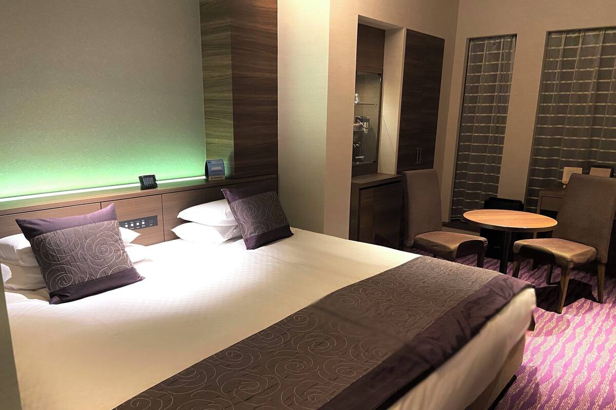 A hotel room with a large bed, two pillows, a bedside table, a chair, a small round table, and a mirror.