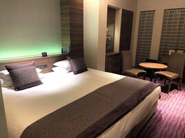 A hotel room with a large bed, two pillows, a bedside table, a chair, a small round table, and a mirror.