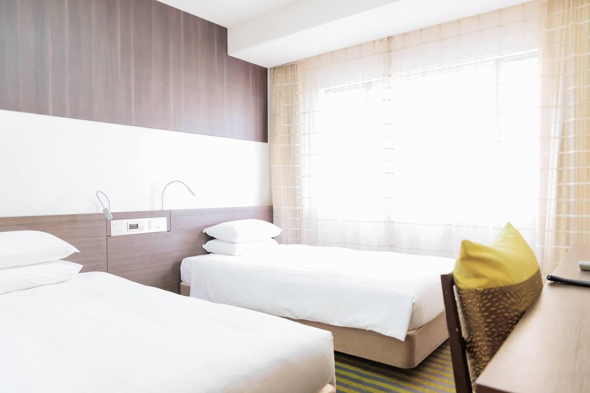 A hotel room with two beds, a bedside table, a chair, and a window with curtains.