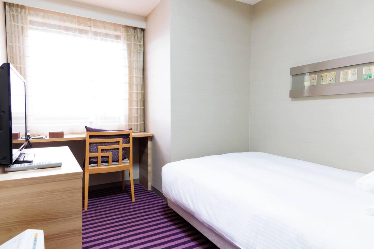 A hotel room with a bed, a desk with a computer, a chair, a window with curtains, and a wall-mounted picture frame.