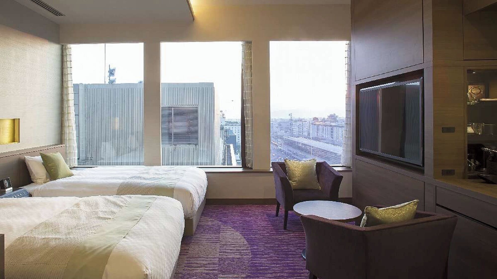 Habitación de hotel con dos camas, televisión, una mesita y vista a la ciudad.