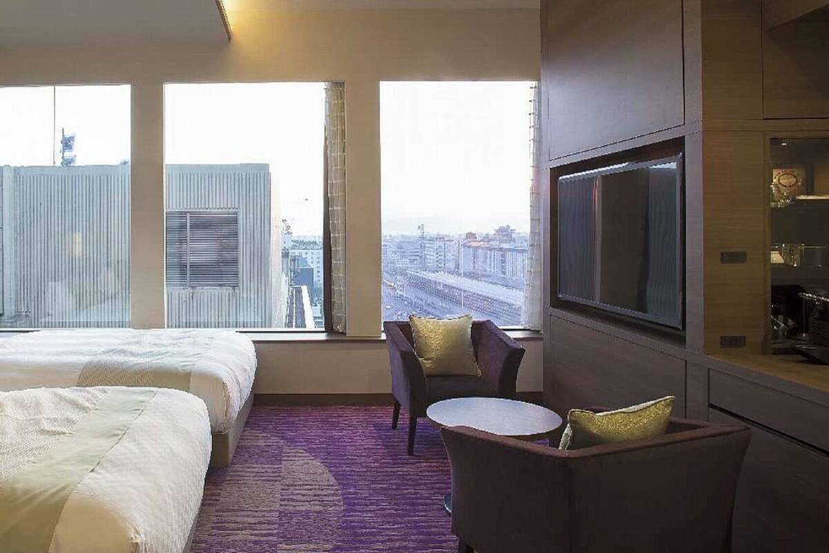 A hotel room with two beds, a TV, a small table, and a view of a cityscape.