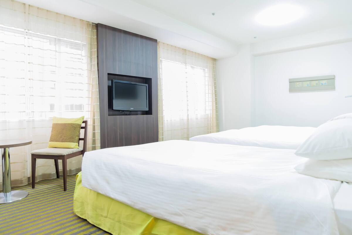 A hotel room with two beds, a TV mounted on the wall, a chair with a cushion, and a small round table.