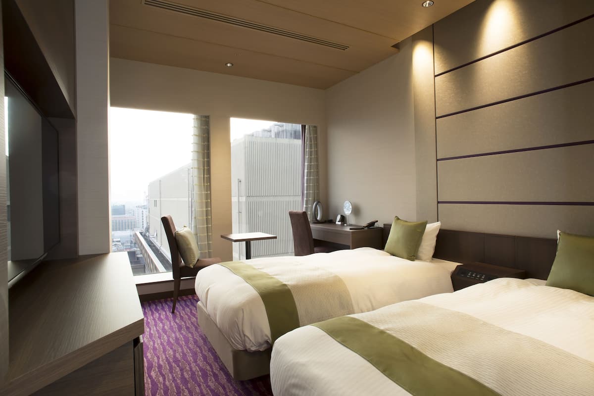 A hotel room with two beds, a desk, a chair, and a view of the city.