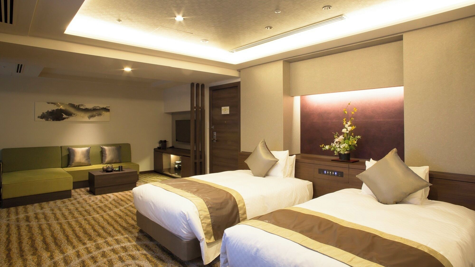 Habitación de hotel con dos camas, un sofá, un televisor, un espejo y una pequeña mesa con un arreglo floral.