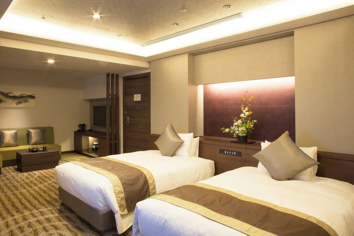 A hotel room with two beds, a sofa, a TV, a mirror, and a small table with a flower arrangement.