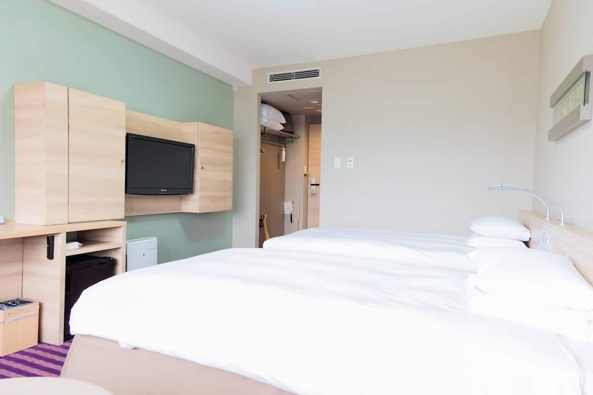 A hotel room with a bed, a TV mounted on a wooden cabinet, a bathroom with a shower, and a small desk with a trash bin.