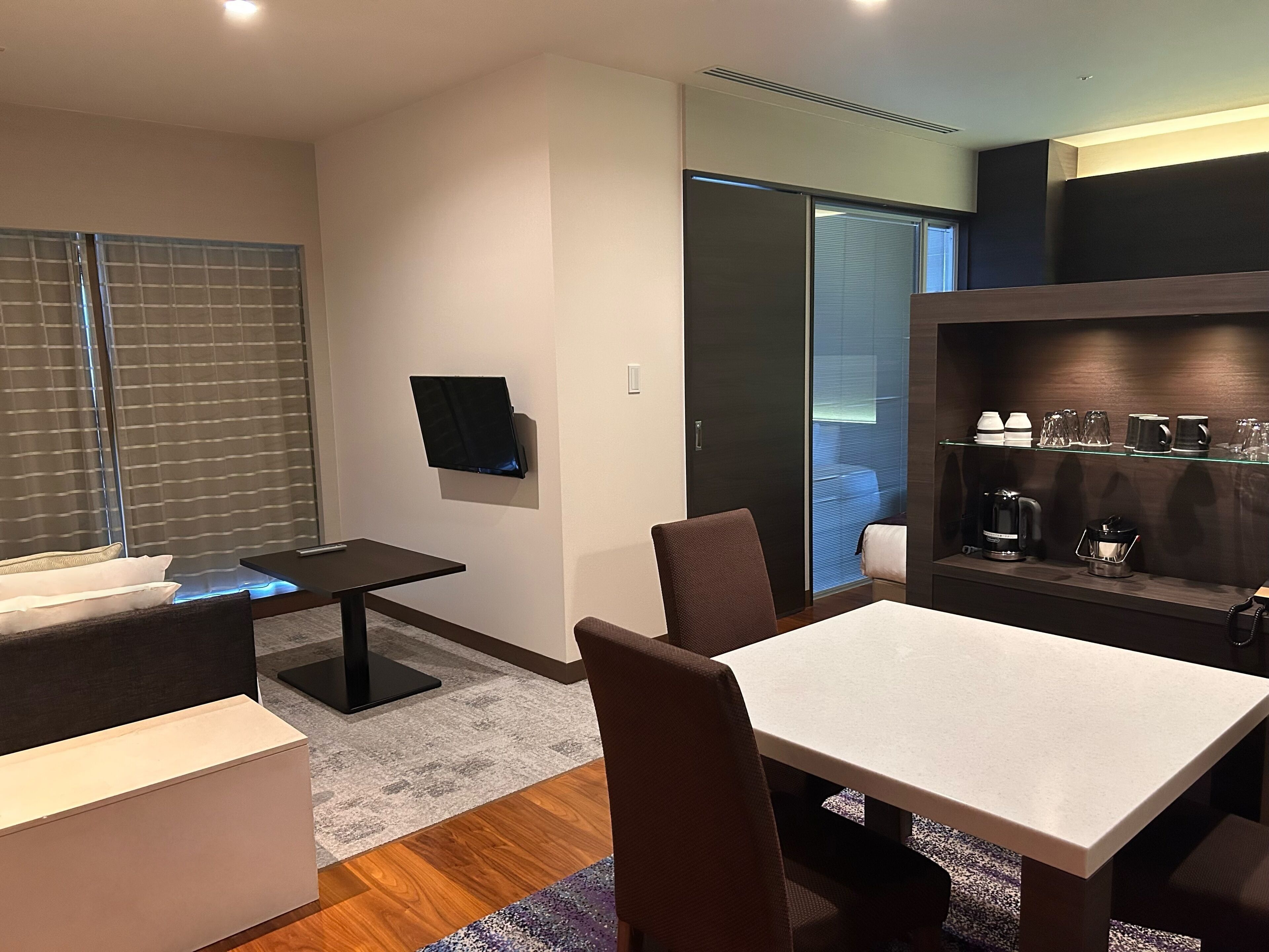Una habitación de hotel moderna con un televisor de pantalla plana montado en la pared, un área de comedor con mesa y sillas, y una kitchenette equipada para preparar café.