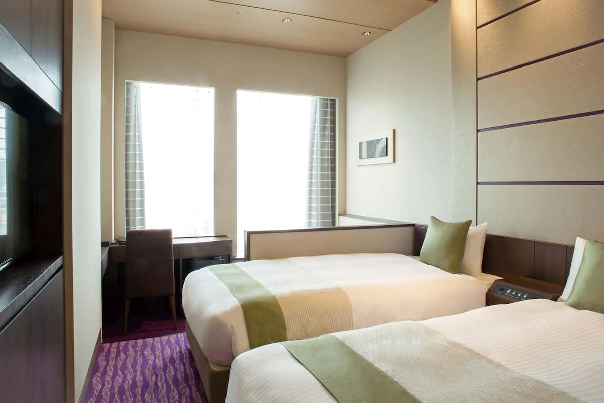A hotel room with two beds, a desk, a chair, and a TV.