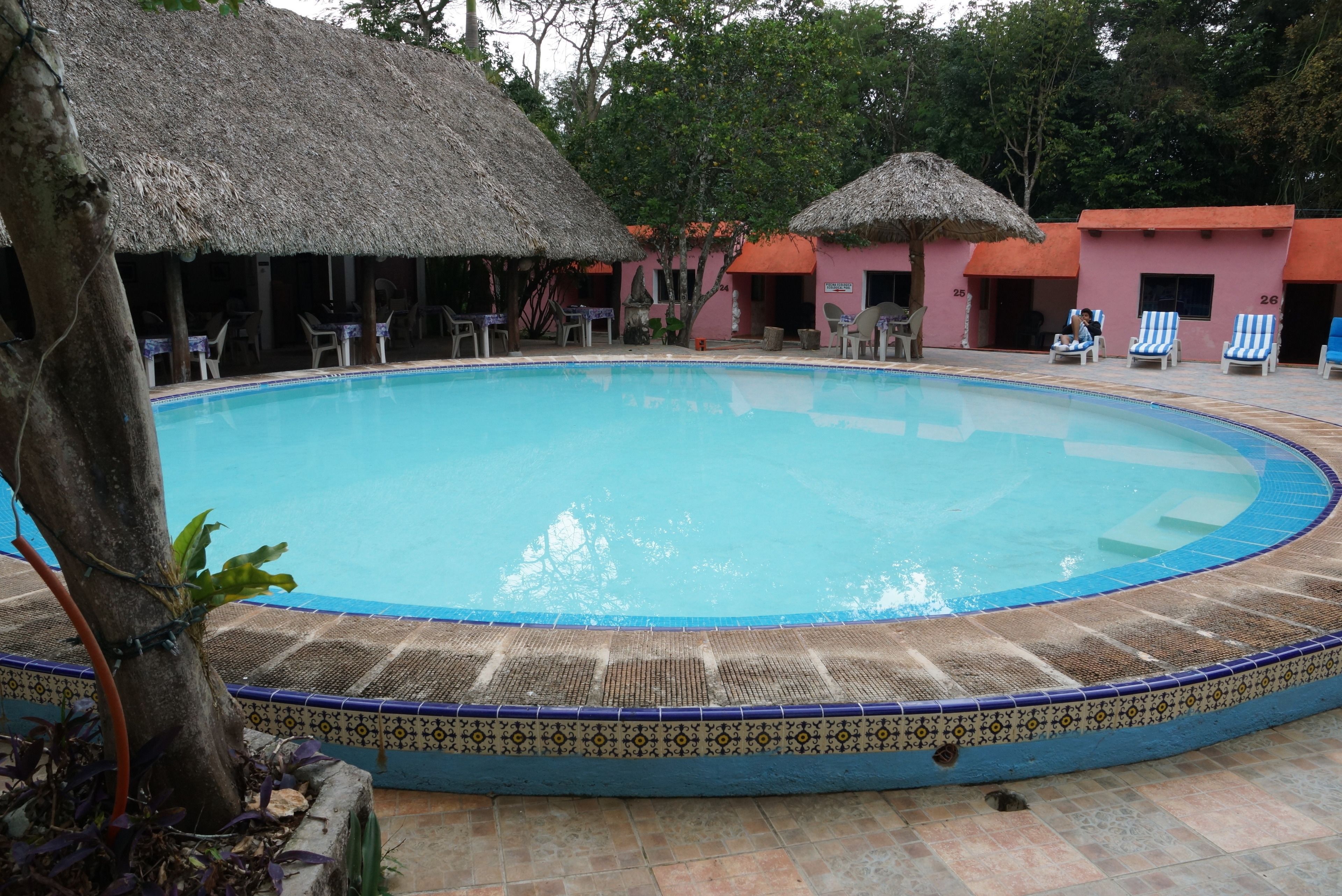 Una piscina con borde de azulejos, rodeada de sillones de descanso y estructuras con techo de paja.
