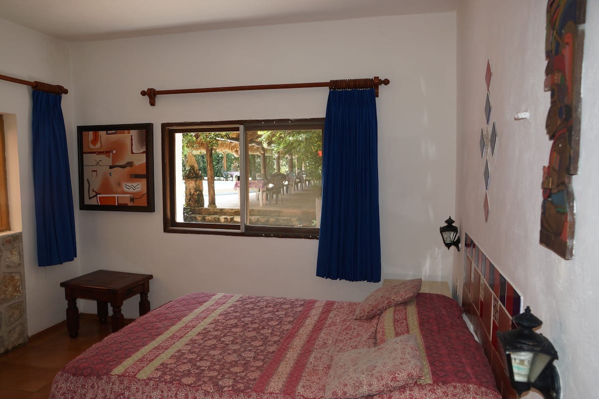 Un dormitorio con cama, ventana con cortinas, una mesita y una lámpara de pared.